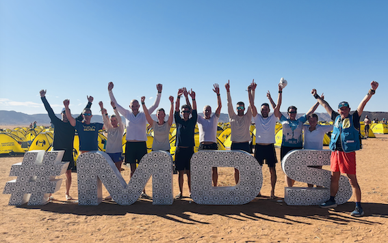 A group of runners with their arms raised in celebration in front of giant #MDS letters at the Marathon des Sables bivouac, yellow tents and Moroccan desert behind them